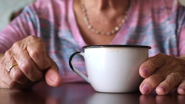 Close up view of an old woman's wrinkled hands with pink nail polish resting on a dark wooden table next to a white enamel mug, showing a concept of waiting, loneliness and aging