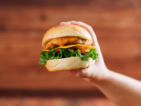 Delicious cheeseburger layer with golden bun juicy meat beef or pork patty melted cheddar cheese holding by hand on wooden background fresh ingredients lettuce tomatoes tasty classic burger junk food.