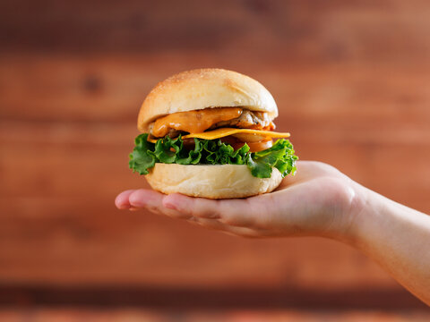 Delicious cheeseburger layer with golden bun juicy meat beef or pork patty melted cheddar cheese holding by hand on wooden background fresh ingredients lettuce tomatoes tasty classic burger junk food.