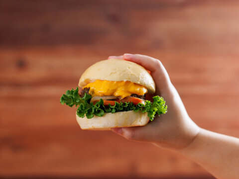 Delicious cheeseburger layer with golden bun juicy meat beef or pork patty melted cheddar cheese holding by hand on wooden background fresh ingredients lettuce tomatoes tasty classic burger junk food.