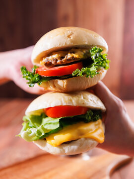 Two delicious homemade cheeseburger stacked together featuring juicy meat beef pork patty melted cheese fresh lettuce tomato holding by hand on wooden background classic burger fast food advertising.