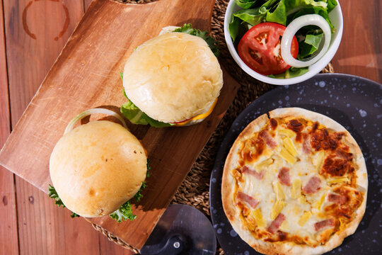 Delicious fresh homemade juicy cheeseburger slice of cheesy pizza over wooden table with fries fresh salad soft drink creating warm appetizing casual dining scene tasty classic fast food advertising.