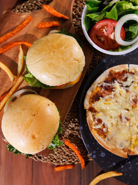 Delicious fresh homemade juicy cheeseburger slice of cheesy pizza over wooden table with fries fresh salad soft drink creating warm appetizing casual dining scene tasty classic fast food advertising.