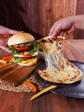 Hands holding delicious homemade cheeseburger slice of cheesy pizza over wooden table with fries fresh salad soft drink creating warm appetizing casual dining scene tasty classic fast food advertising
