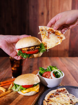 Hands holding delicious homemade cheeseburger slice of cheesy pizza over wooden table with fries fresh salad soft drink creating warm appetizing casual dining scene tasty classic fast food advertising