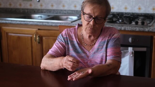 Senior woman with glasses sitting at her kitchen table, carefully counting and taking her daily dose of medication for health treatment, highlighting the routine of elder care and healthcare