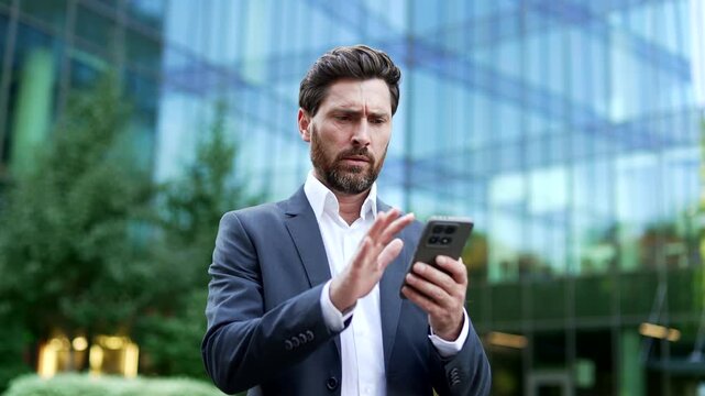 Annoyed manager in formal suit reacting with irritation to payment error on mobile phone outside modern glass office building. Frustrated businessman facing failed transaction declined on app.
