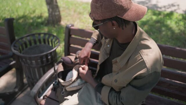 Close up of man sitting on bench in park rummaging through bag. Man wearing cap and jacket interacts with items inside handbag while seated outdoors near trash can. Bright daylight