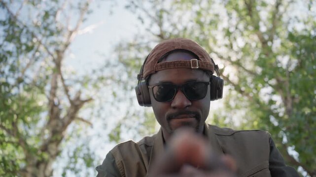 Close up of man wearing sunglasses and headphones gesturing with his hands in outdoor setting. subject is dressed in casual attire including cap and jacket interacting directly towards