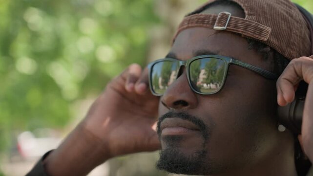 Close up of man wearing sunglasses and ear protection adjusting headphones in outdoor park setting with green foliage. subject has braided hair under brown cap and is seen from various