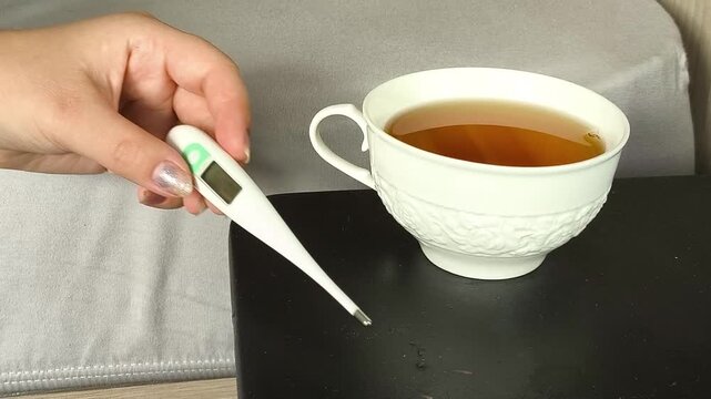 Woman placing a thermometer on a table after checking her temperature and picking up a cup of tea, cold and flu, fever and home treatment concept. flu season