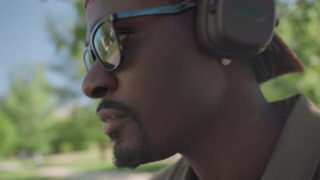 Close up of man wearing brown cap and ear protection in park setting with green trees background. Man remains stationary fine details include sunglasses reflection showing outdoor scene