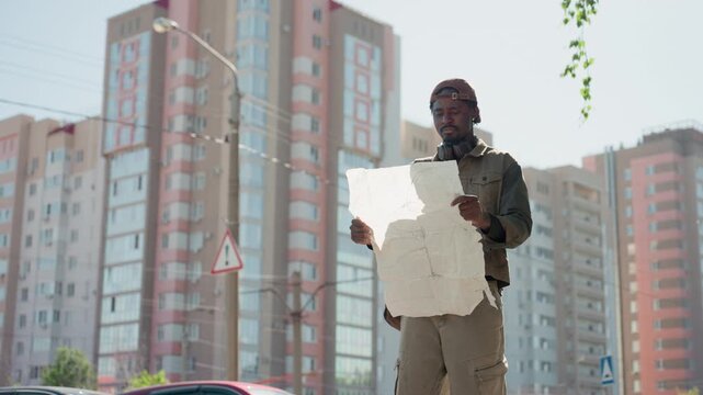 Man in work attire examines large white paper map or plan outdoors against urban backdrop of tall residential buildings under bright daylight. Man holds and unfolds document revealing