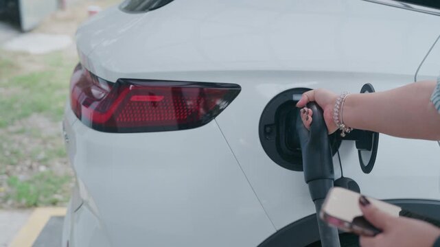 Slow motion close up of woman plugging charging cable into white electric vehicle while holding smartphone.