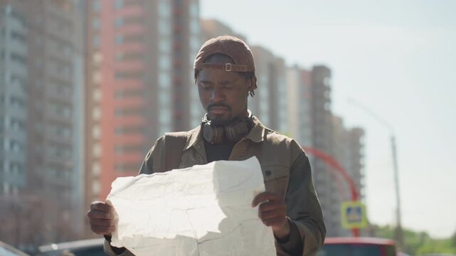 Close up of man holding and examining large white paper map outdoors with trees cars buildings in background. Man wears brown cap jacket headphones around neck carries backpack. Paper shows