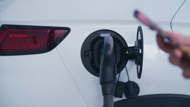 Slow motion close up of woman plugging charging cable into white electric vehicle while holding smartphone.