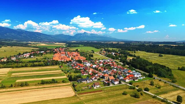 Aerial view of Slovak village with High Tatras mountains background. Rural settlement with red roofs surrounded by green fields, forest, High Tatras mountain ridge under blue sky