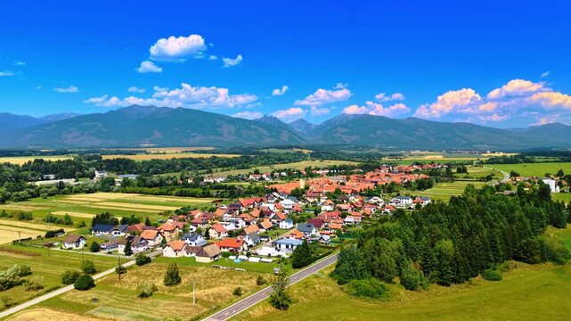 Village near green forest and fields with High Tatras mountain range. Aerial view of countryside village bordered by dense green pine forest, fields, road, High Tatras mountains under blue sky