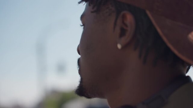 Close up of man with braided hair and earring wearing casual clothing. background consistently shows urban elements like traffic lights on poles under clear daylight sky. Soft focus effect
