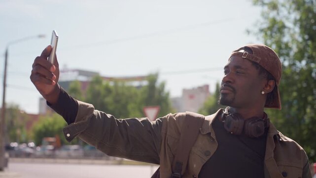 Close up of man taking selfie outdoors with smartphone. Man wears brown cap jacket and headphones around neck in urban setting with trees and buildings visible. Soft daylight illuminates