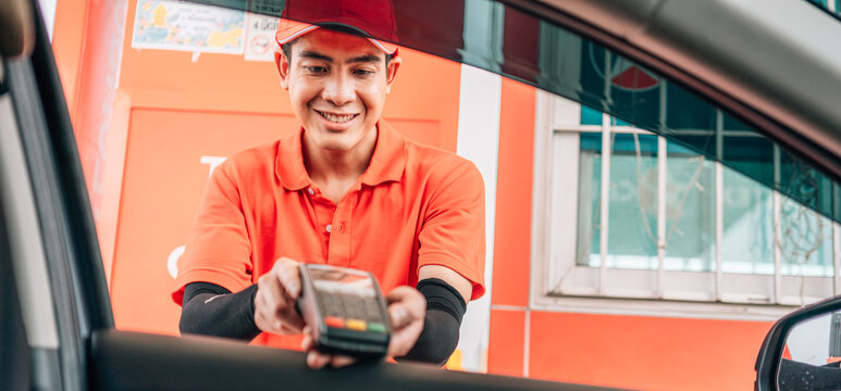 Smiling gas station attendant offering a wireless POS terminal through a car window for cashless payment.