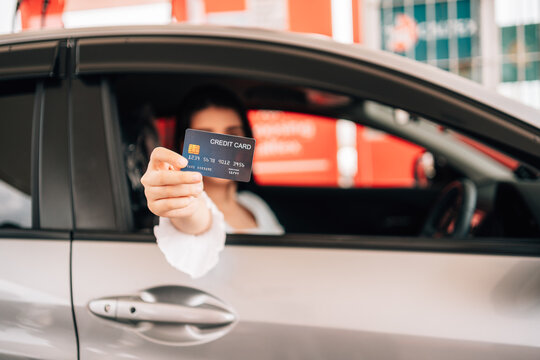 A driver's hand extends a dark credit card directly toward the camera through an open car window, with the woman in the driver's seat softly blurred in the background