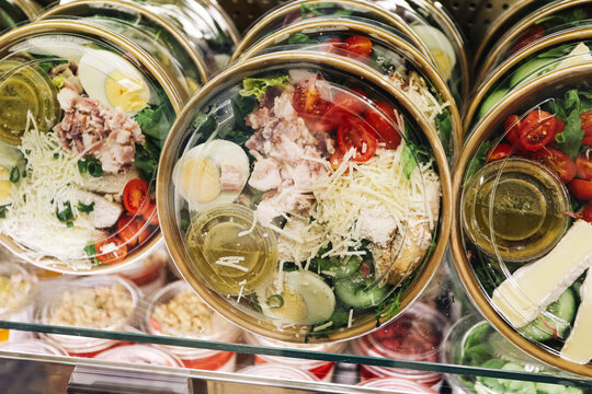 Fresh prepared salads in transparent takeaway bowls inside refrigerated display. Vegetables eggs cheese and meat form healthy food background. Ready meals for quick lunch and retail sale.