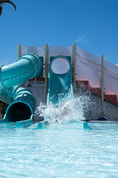 Bright blue slide plunges into sparkling pool. Splash erupts as child emerges from tube ride. Joyful laughter echoes under clear sky. Ideal for water park, summer fun, or family vacation themes