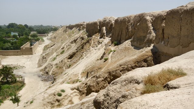 Ancient walls of Balkh in northern Afghanistan. Eroded mudbrick cliffs with dry riverbed and greenery under clear sky