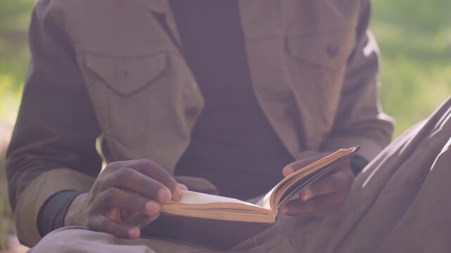 Close up of man reading open book while seated outdoors in natural light. man holds pages with both hands focusing on text throughout scene. Soft sunlight illuminates details like page