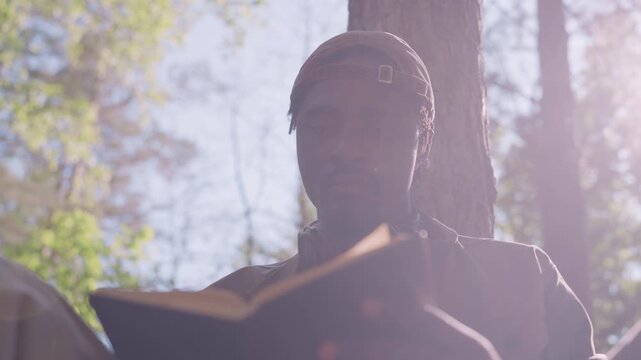 Close up of man reading open book outdoors with sunlight filtering through trees. subject holds pages steady focusing on text in natural daylight surrounded by green foliage and tree trunks