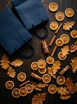 Navy gift bags surrounded by dried orange slices, cinnamon sticks, star anise and autumn leaves on a dark wooden table, a festive flat lay.
