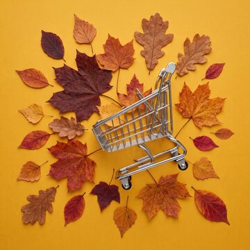 A small metal shopping cart sits on a bed of colorful autumn leaves against a yellow background, an autumn sale and shopping concept.