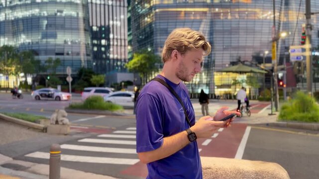 Young Blond Man Standing at Pedestrian Crossing in Seoul at Night Using Smartphone &ndash; Urban Street Scene with City Lights