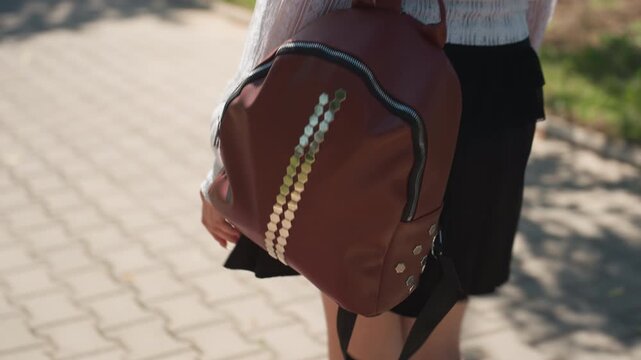 Close up of young woman carrying brown backpack with gold studs walking outdoors on paved ground. subject wears white top and black skirt focusing solely on lower torso and bag details. Soft