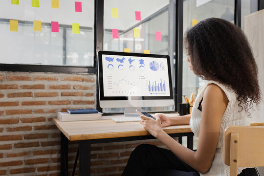A young African American businesswoman working on a computer with financial data charts on the screen in a modern office.