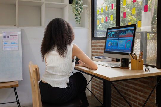 A young African American businesswoman working on a computer with financial data charts on the screen in a modern office.
