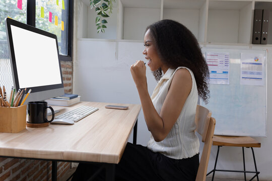 A joyful African American businesswoman celebrating a successful outcome while working at her desk with a computer surrounded by a modern office workspace.