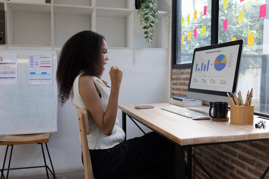 A happy African American businesswoman celebrating success while looking at financial charts and graphs on her computer monitor in a modern office.
