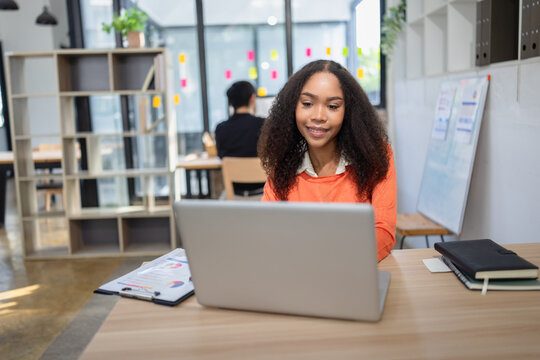 A smiling African American businesswoman holding a notebook and pen at her modern office desk, with a colleague working in the background.