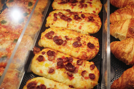 Cheese topped bakery snacks with sliced sausage beside croissants behind display glass. Golden baked pastry assortment forms savory fast food bakery counter background for retail sale.
