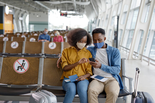 A man and woman wearing face masks sit together in an airport departure lounge, checking their mobile phone and passports