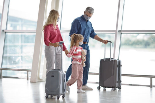 A family of three wearing protective face masks walks through a modern airport terminal with their suitcases and travel documents
