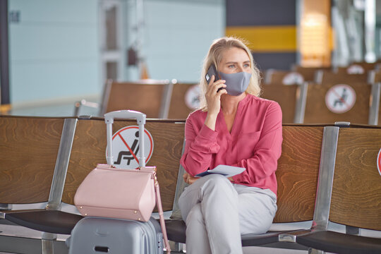 A woman in a face covering speaks on her mobile phone while seated in an airport waiting area with signs indicating social distancing measures