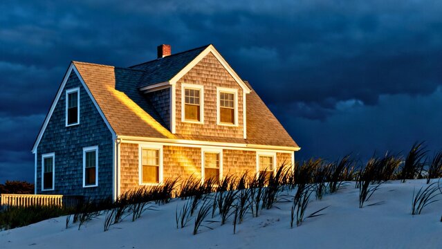 A crisp grey-shingled Cape Cod cottage with bright white trim against a deep navy storm-clearing sky, low golden-hour sun creating strong shadows and texture.