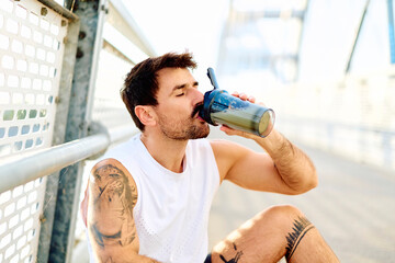 Fototapeta premium Male athlete hydrating with a protein shake on a bridge, recovering after an outdoor training session for fitness and wellness