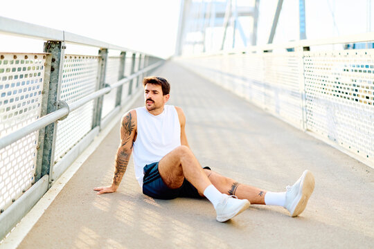 Male runner with tattoos sitting on a modern bridge, looking away after a strenuous urban workout, feeling tired and exhausted