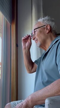 Pensive older man with a goatee and gray hair sitting in a chair, thoughtfully putting on his glasses while looking out the window, conveying a sense of loneliness and reflection