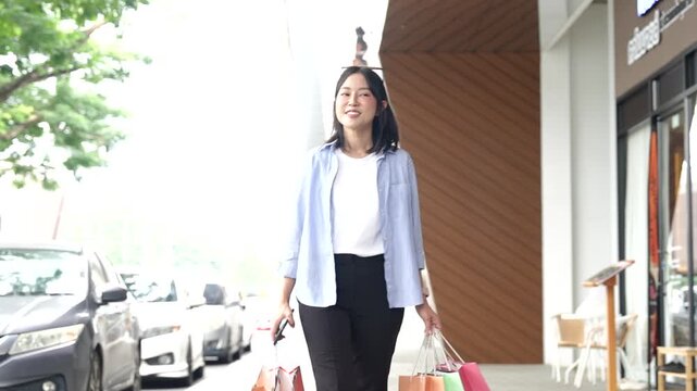 Asian woman happily carrying colorful shopping bags outdoors near a modern shopping center, representing retail, fashion, consumerism, shopping lifestyle, and urban leisure.