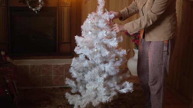 Woman decorating white Christmas tree with Christmas ornaments in cozy home.